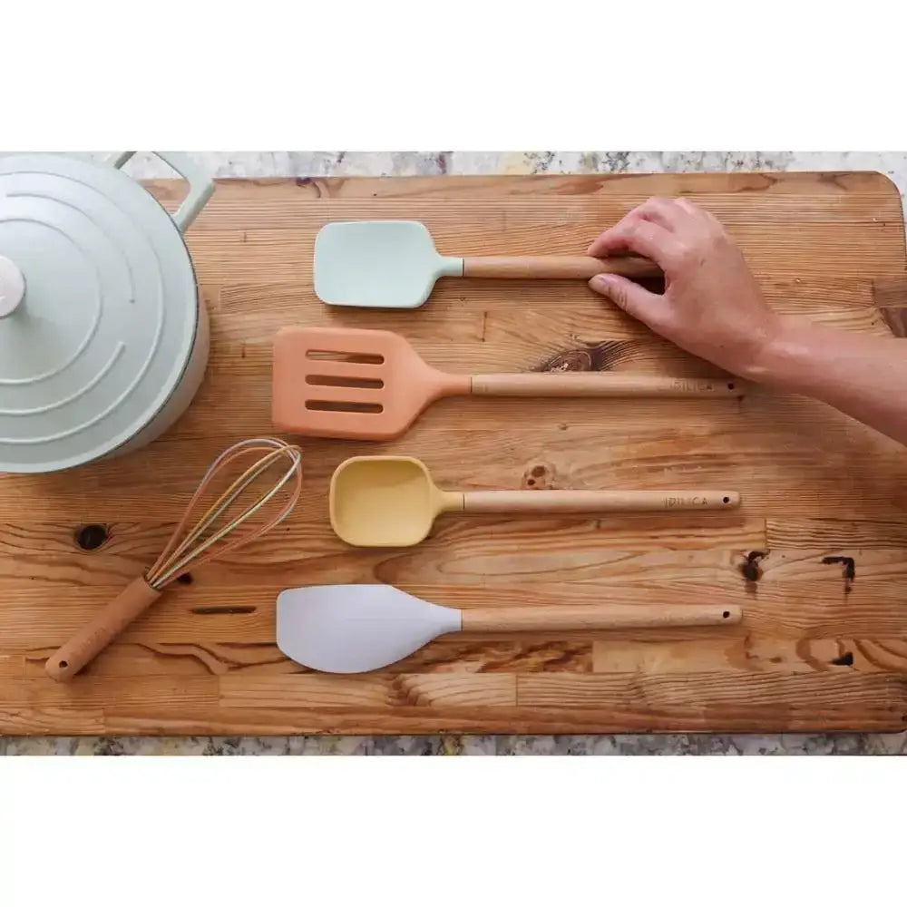 Assorted silicone kitchen utensils on wooden board next to Dutch oven, hand arranging spatula