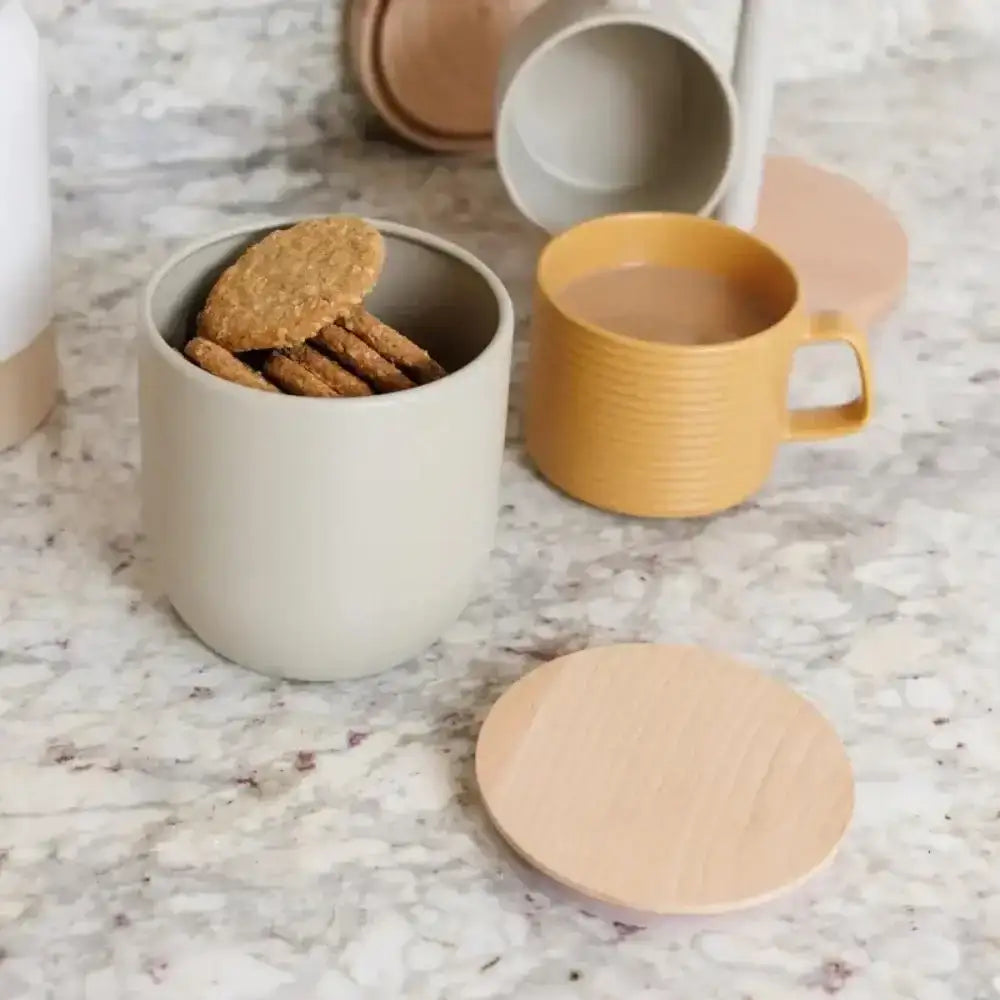 Storage jar with cookies, yellow mug with liquid, and coasters on a marble surface