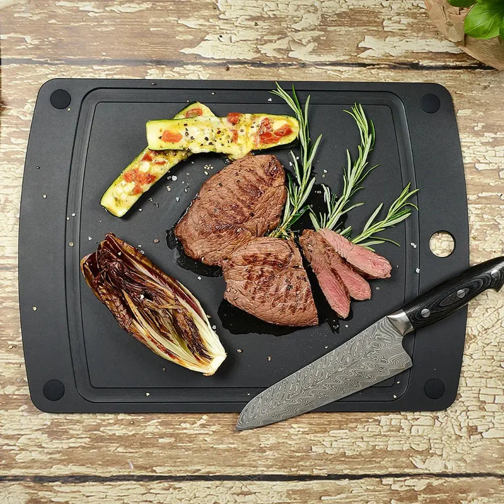 Sliced steak on a black cutting board with vegetables and a knife on a wooden surface