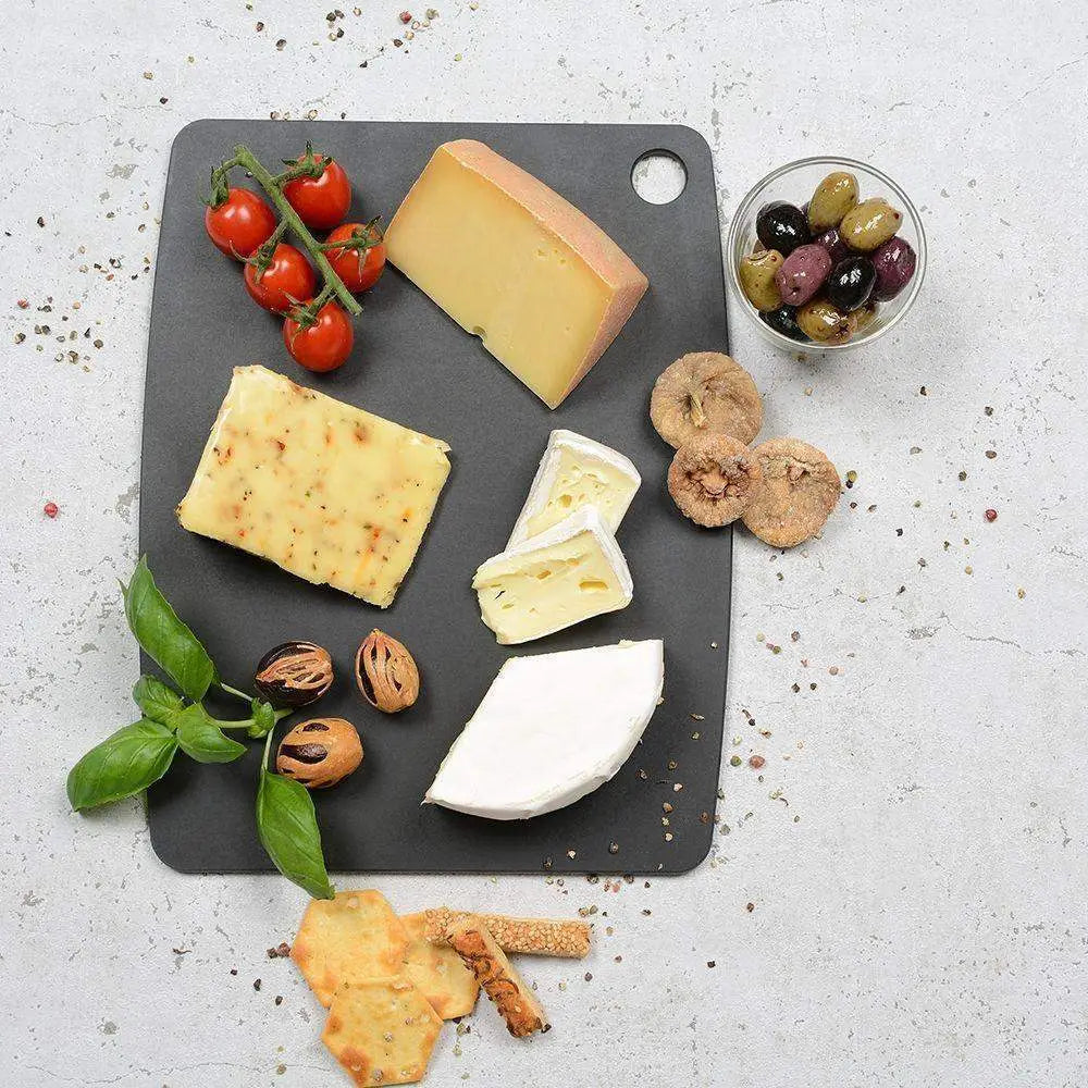 Assorted cheeses on a dark slate board with crackers, grapes, and herbs on a light gray background