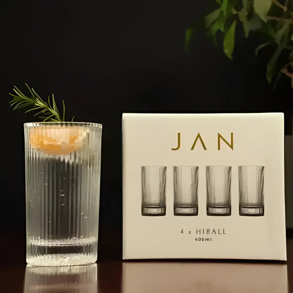 Clear glass with a drink and rosemary garnish next to a box of 'JAN' glasses on a dark surface.