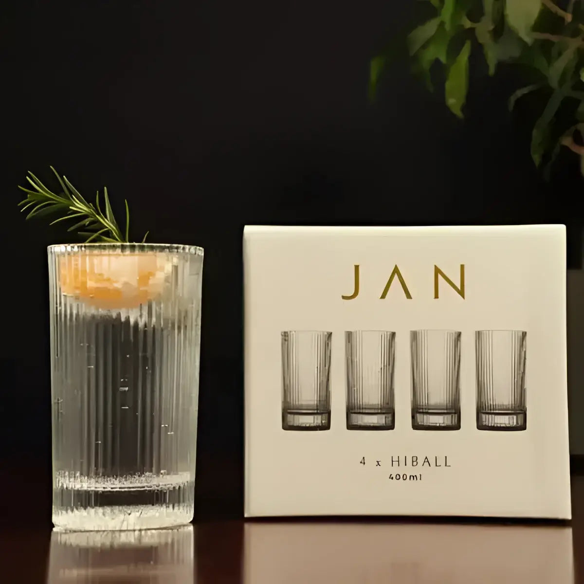 Clear glass with a drink and rosemary garnish next to a box of 'JAN' glasses on a dark surface.