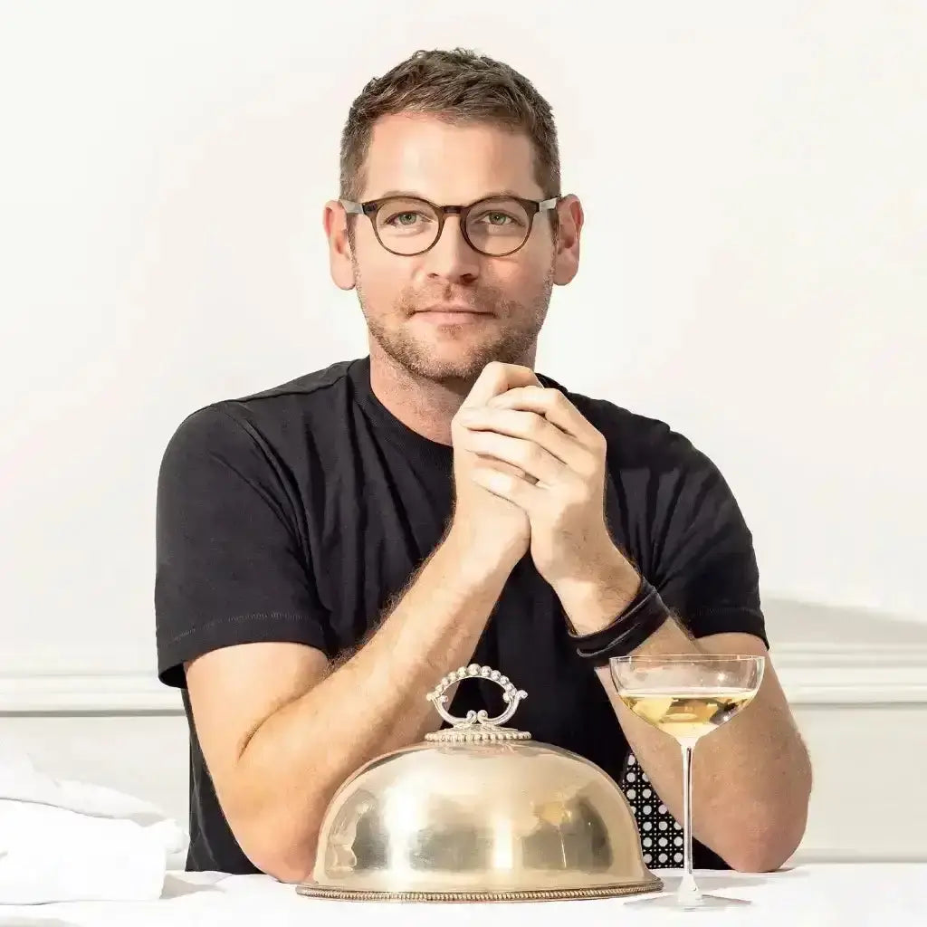 JAN Hendrick sitting at a table with a glass of wine and a covered dish on a white background