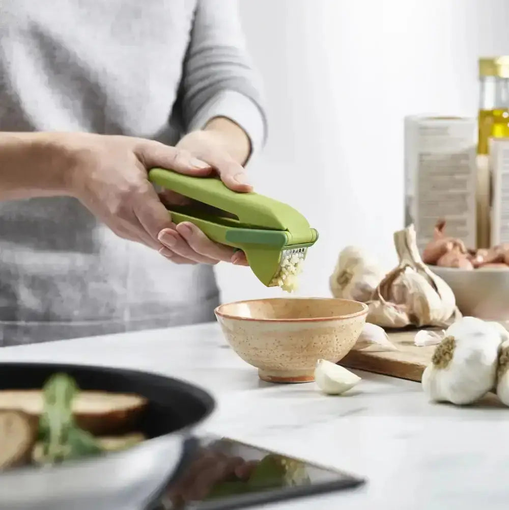 Person using a green garlic press on a kitchen counter with garlic and a bowl.