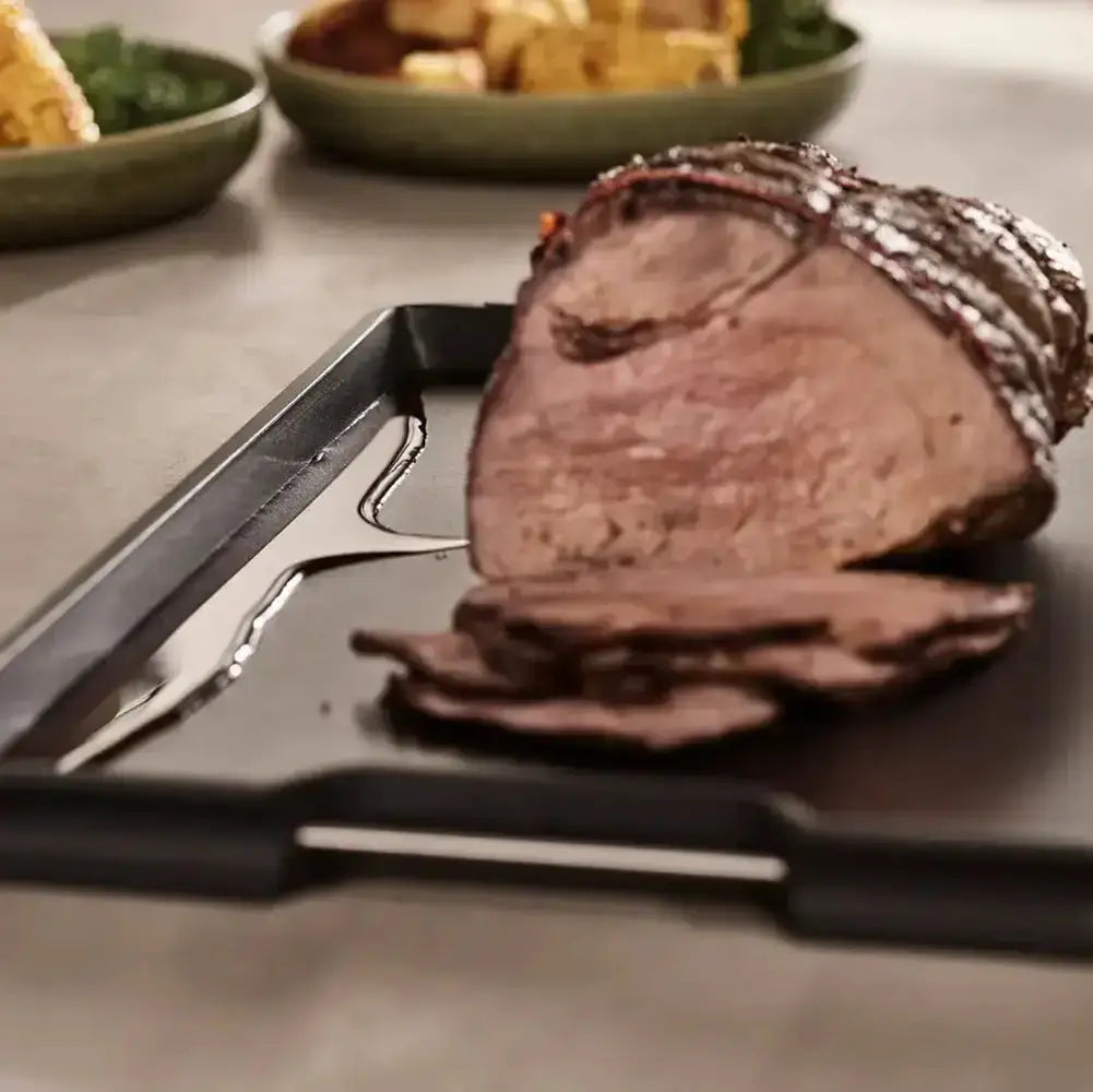 Sliced roast beef on a cutting board with a knife and fork, surrounded by other dishes.