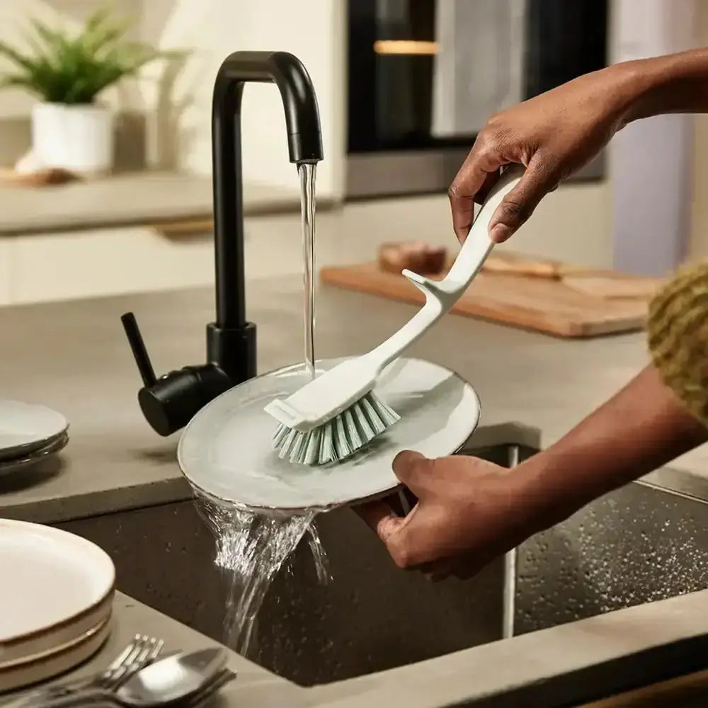 Person washing dishes in a kitchen sink with running water and a dish brush.