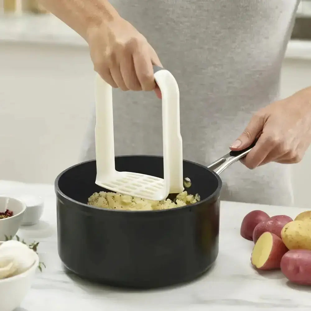 Person mashing potatoes in a pot with a white masher, surrounded by ingredients on a kitchen counter.