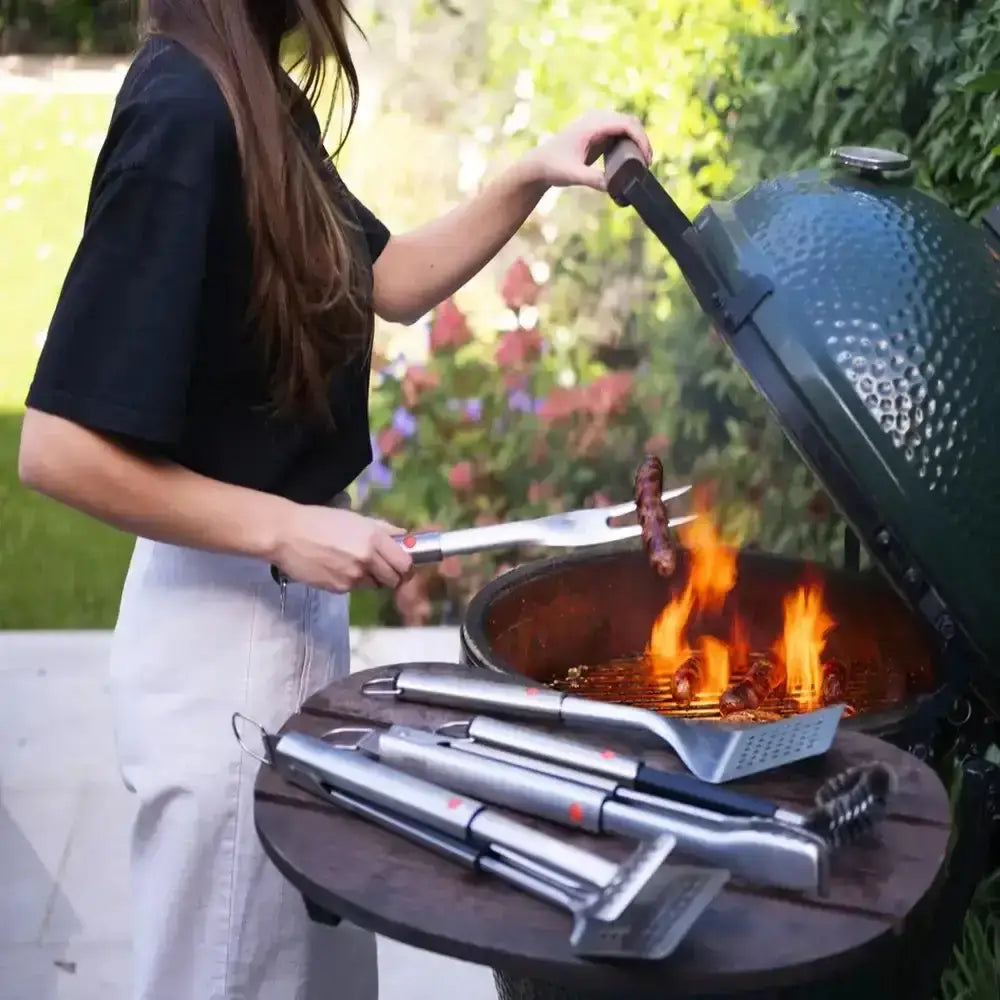 Person grilling outdoors with a green barbecue and various grill tools on a wooden table.