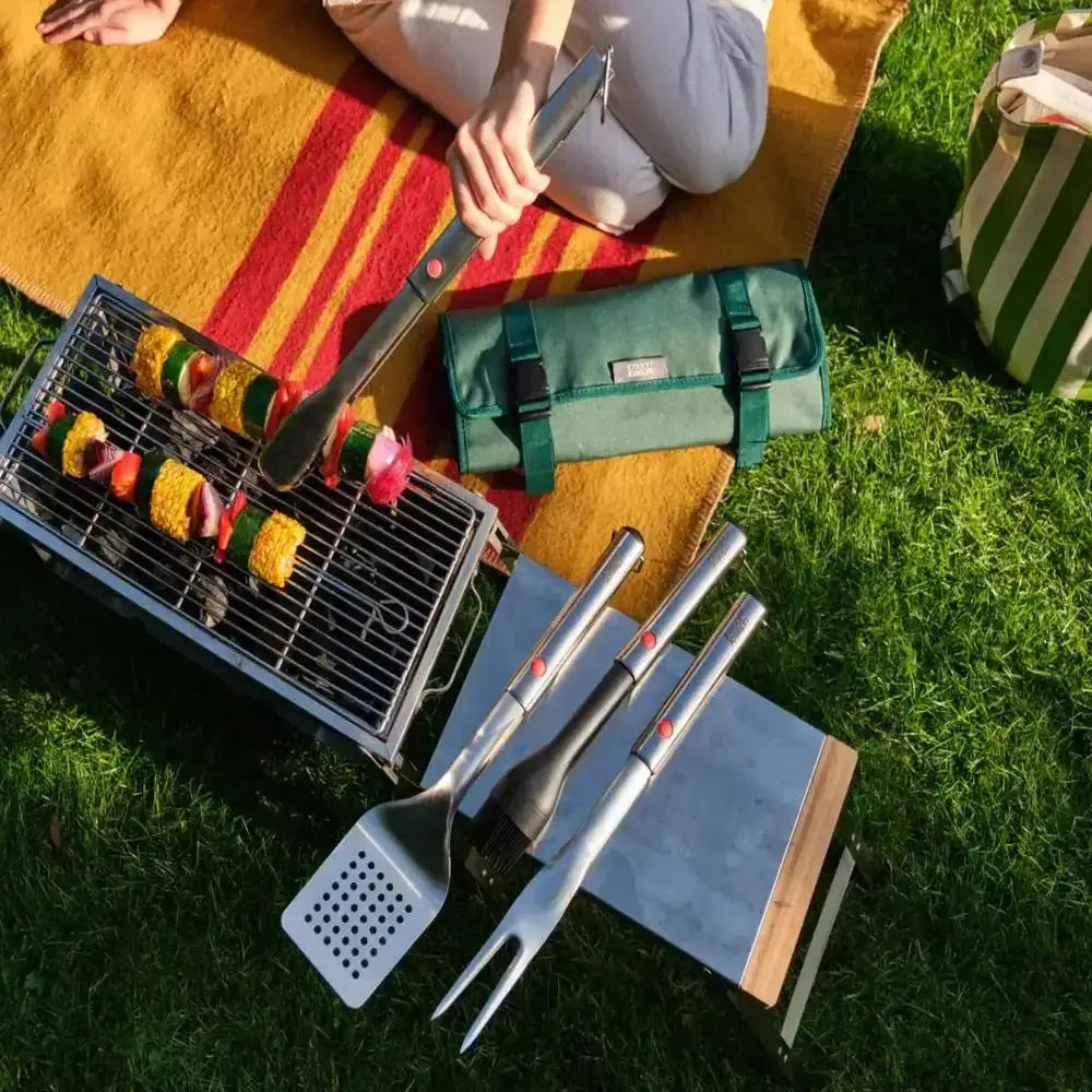Grill with skewered food, grill tools, and a green bag on a grassy area.
