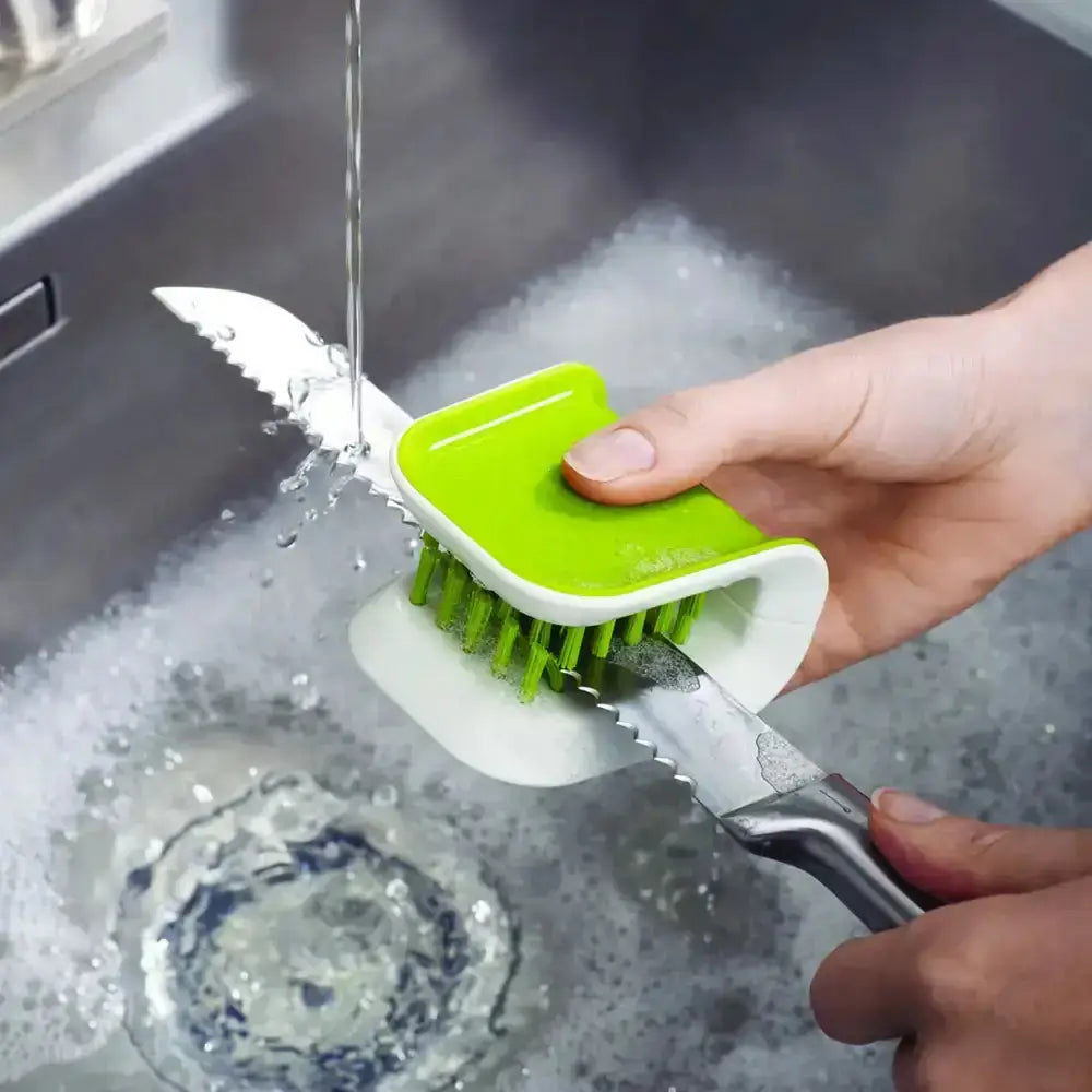 Person using a green kitchen brush to clean a knife under running water in a sink.