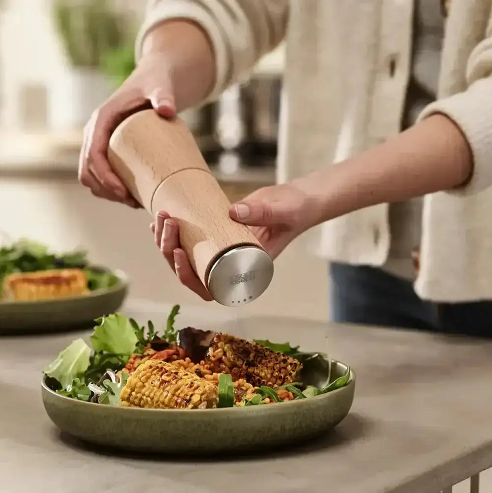 Person seasoning a salad with a wooden pepper grinder in a kitchen setting