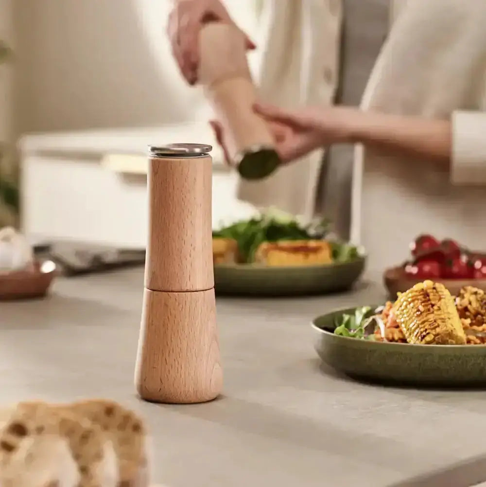 Wooden pepper grinder on a kitchen counter with food in the background
