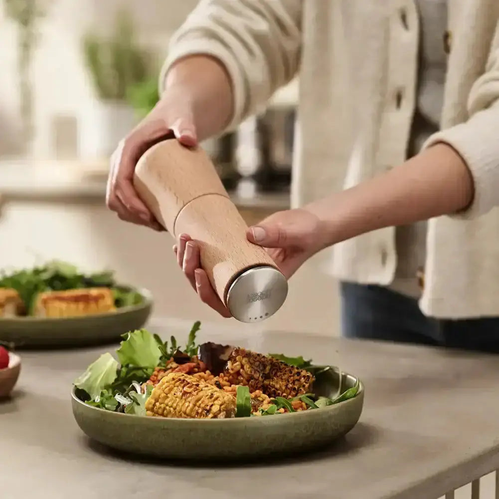 Person seasoning a salad with a wooden salt grinder in a kitchen setting.