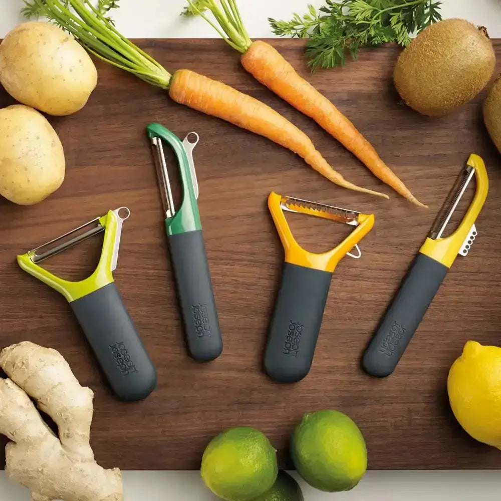 Three vegetable peelers on a wooden cutting board with vegetables around