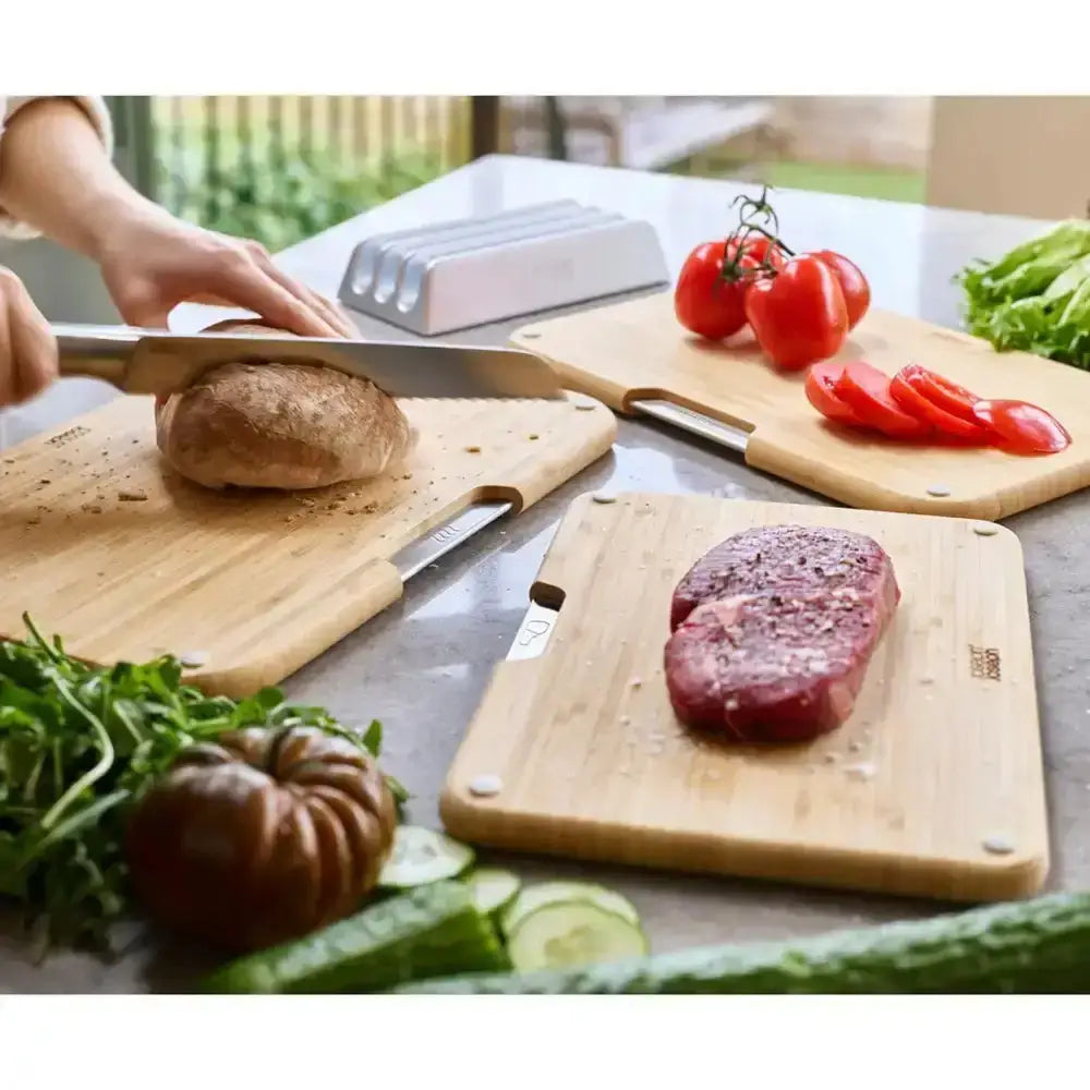 Person slicing bread on a wooden cutting board with vegetables and a steak in the background.