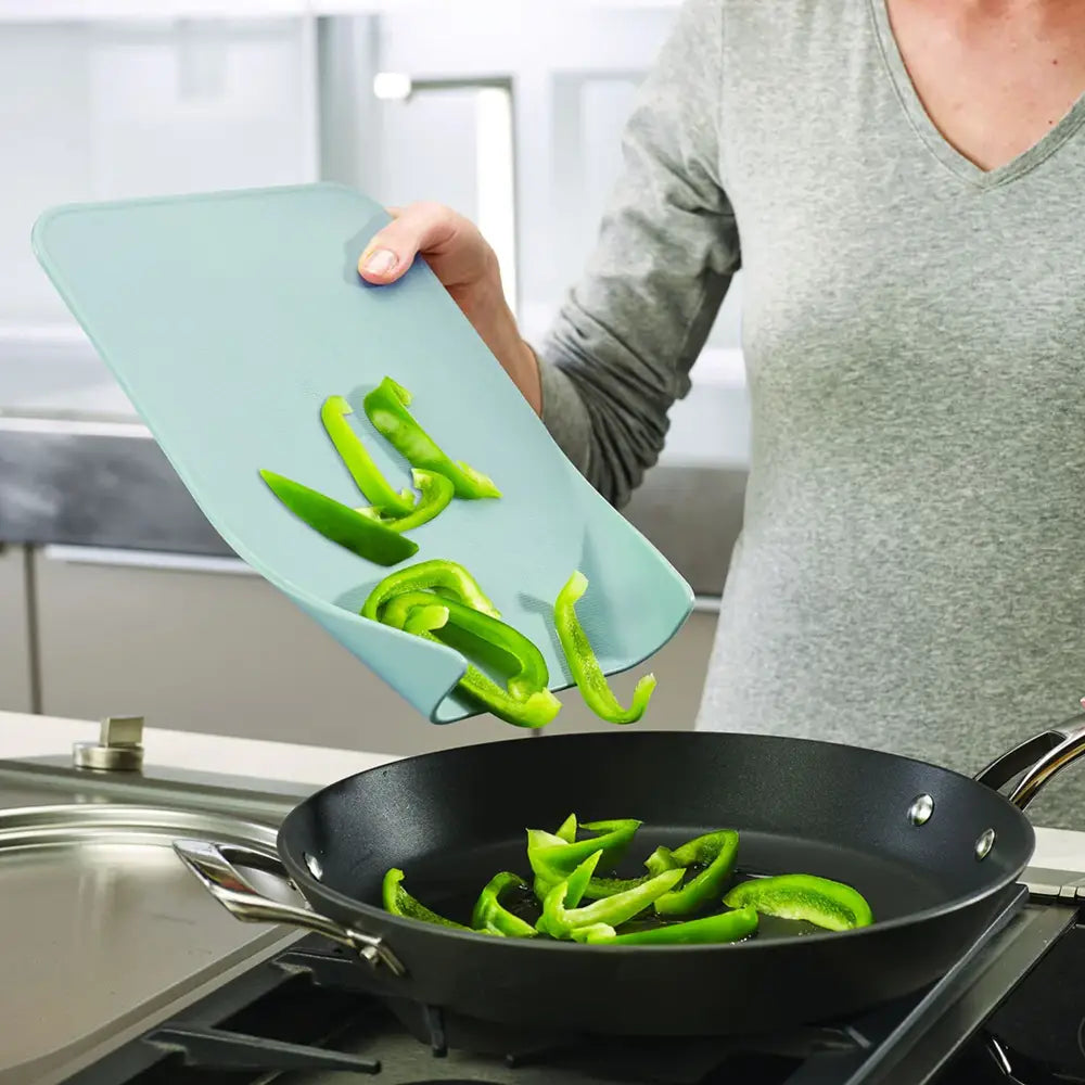 Person transferring green bell peppers from a cutting board to a frying pan in a kitchen.