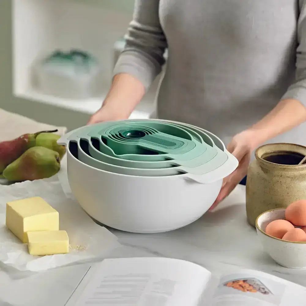 Stack of white and green nesting bowls on a kitchen counter with a person and various items in the background.
