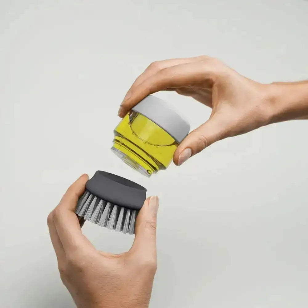 Person holding a gray scrub brush and a clear container with a yellow liquid on a light gray background