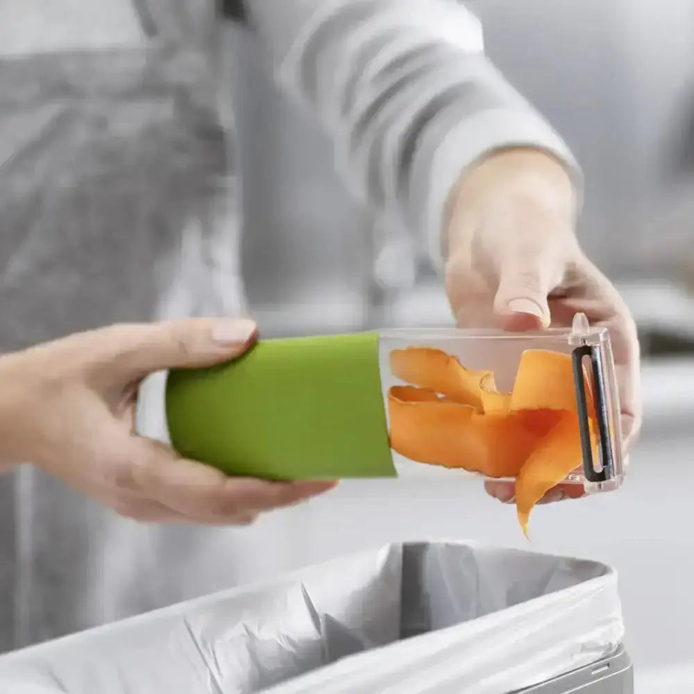 Person using a vegetable peeler to remove orange skin from a potato over a trash bin.
