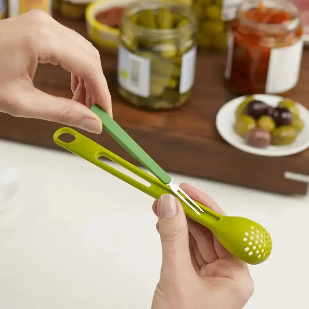 Green olive pitter held by a hand with jars of olives in the background