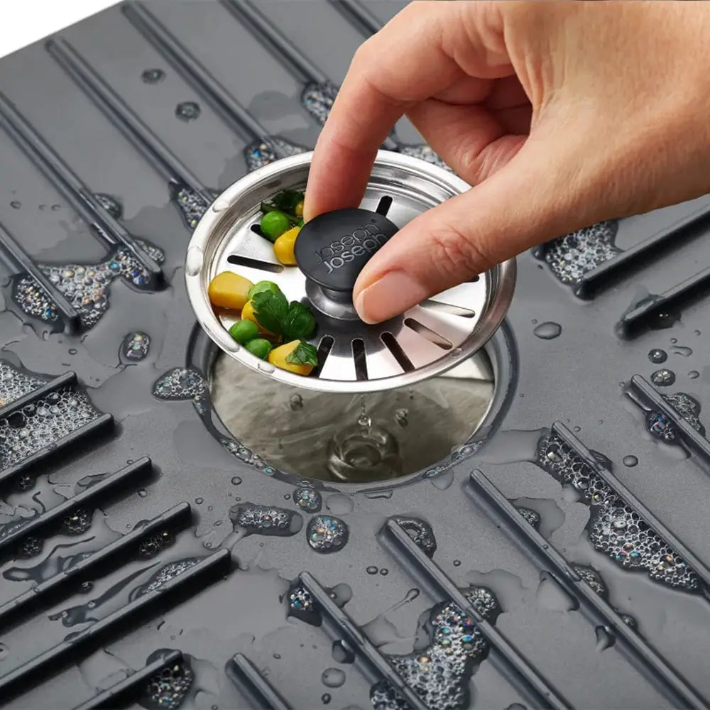 Hand placing a black sink stopper into a silver drain with vegetables on a gray surface.