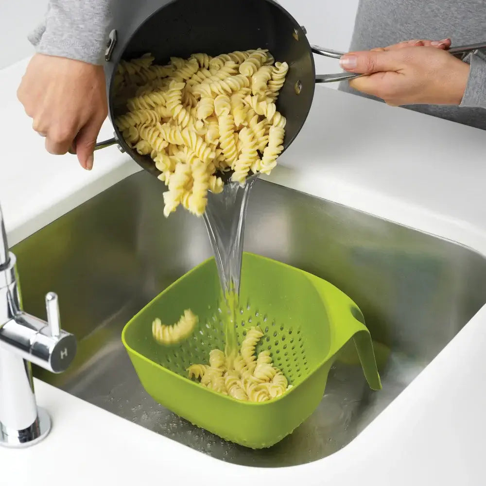 Pasta being drained into a green colander in a kitchen sink.