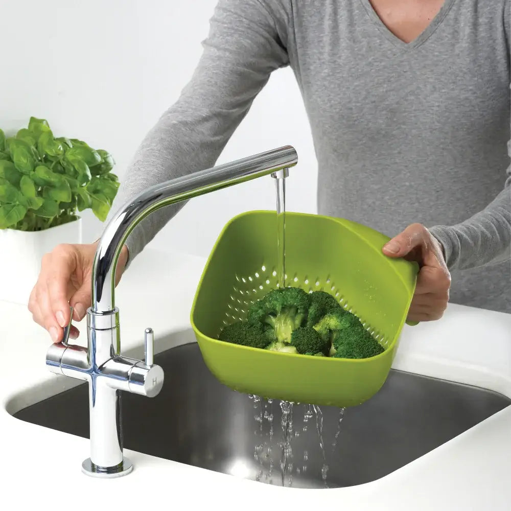 Person washing broccoli in a green colander over a sink