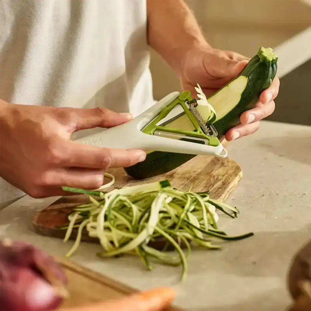 Person using a vegetable peeler to slice a zucchini on a wooden cutting board.