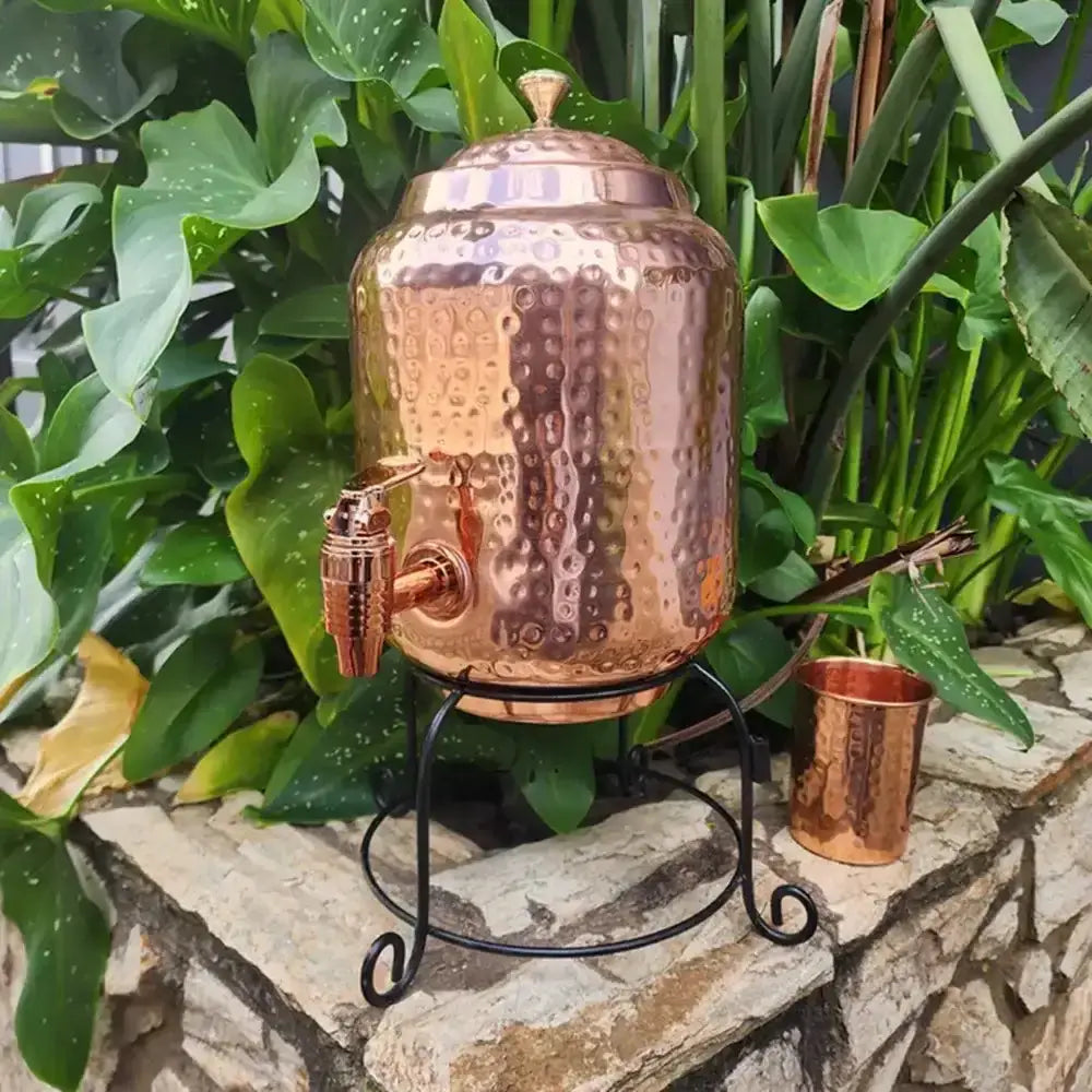 Copper dispenser with a cup on a stone surface with green plants in the background
