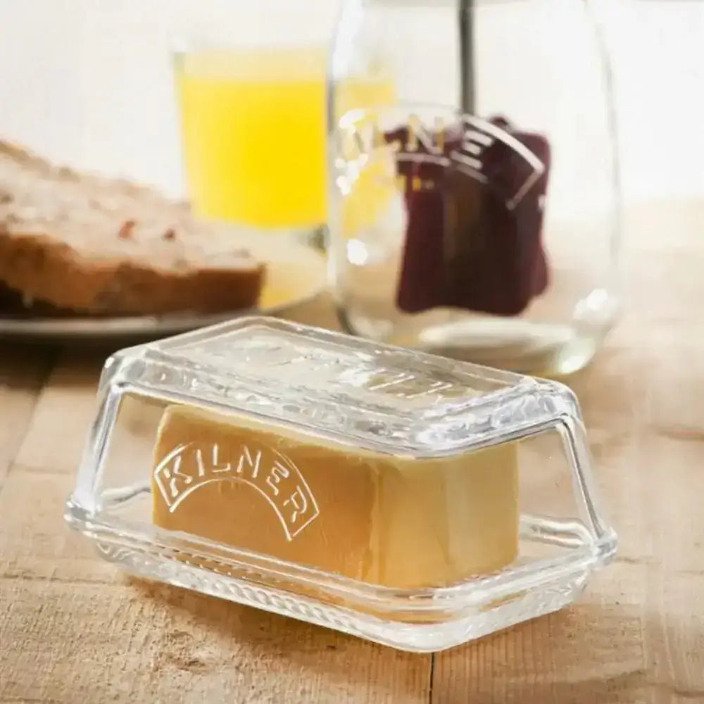 Glass butter dish with stick of butter on wooden table, bread and juice in background