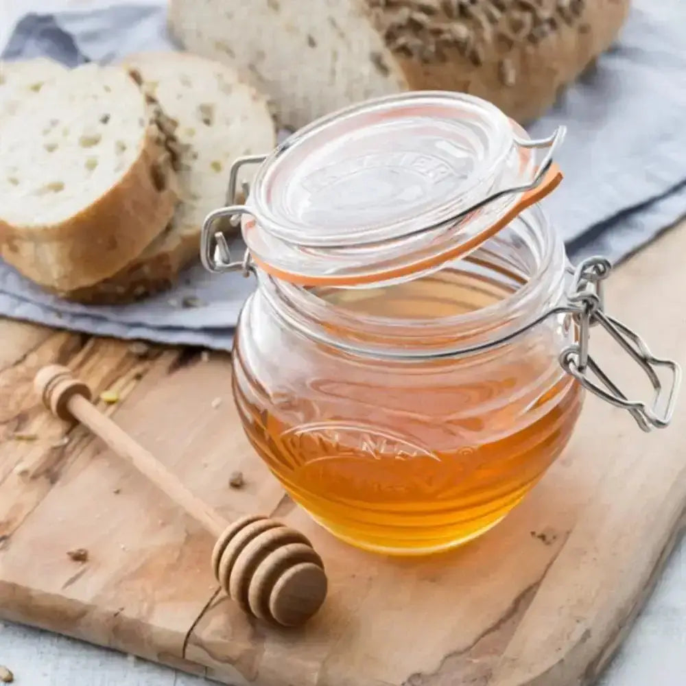 Jar of honey with a wooden honey dipper on a wooden board with bread slices in the background.