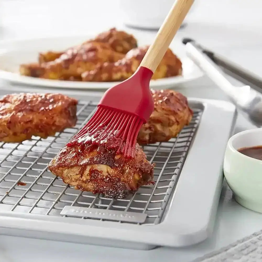 Red silicone brush applying sauce to chicken wings on a cooling rack with a blurred background.