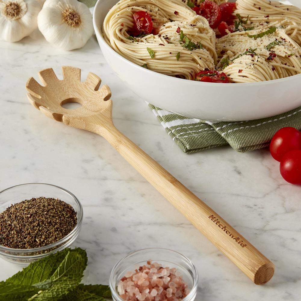 Wooden pasta server with 'KitchenAid' branding on a marble surface with a bowl of pasta and ingredients.