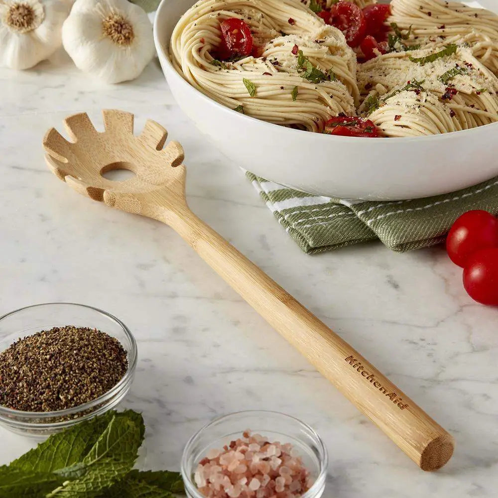 Wooden pasta server with 'KitchenAid' branding on a marble surface with a bowl of pasta and ingredients.