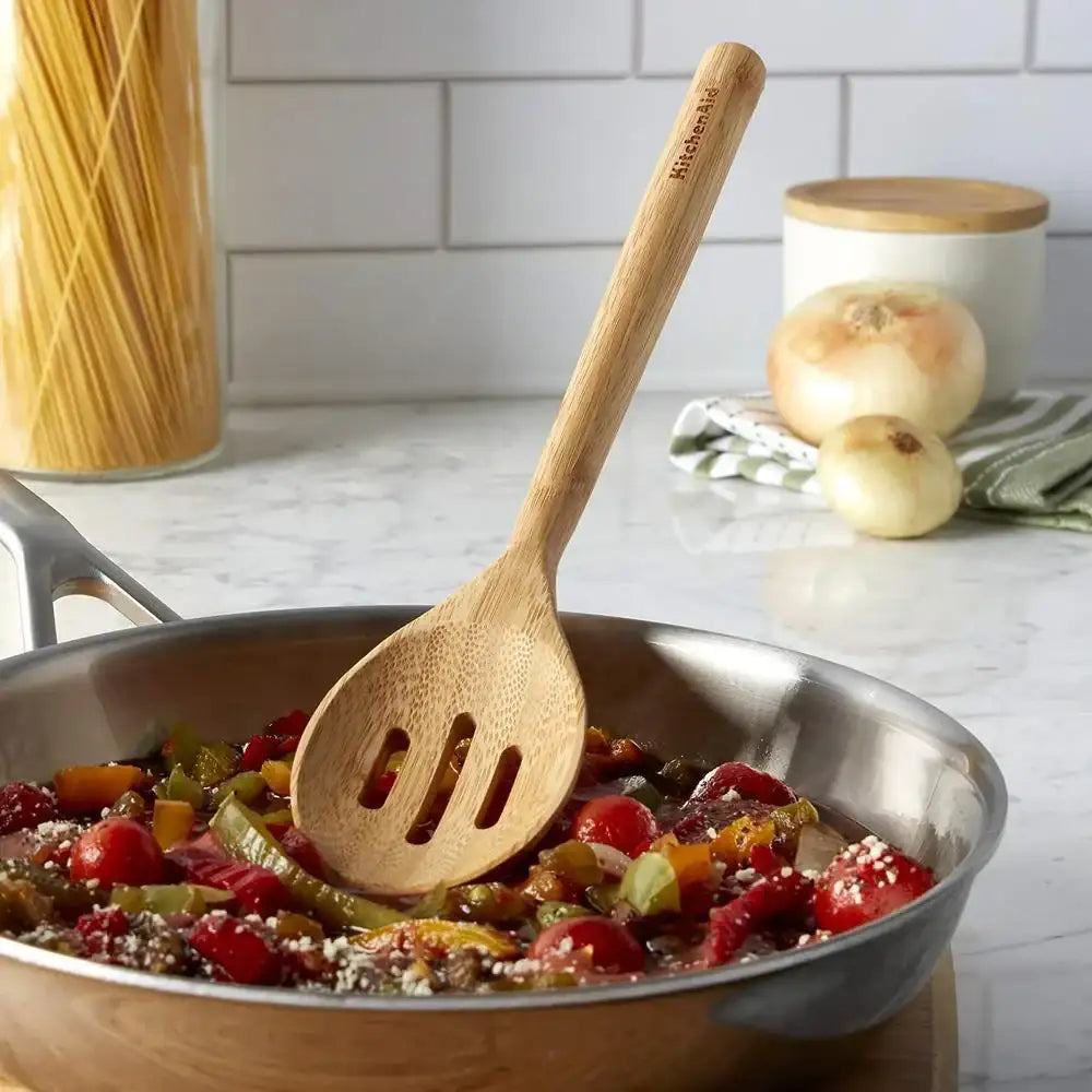 Wooden slotted spoon in a pan of food on a kitchen counter with onions and pasta in the background.