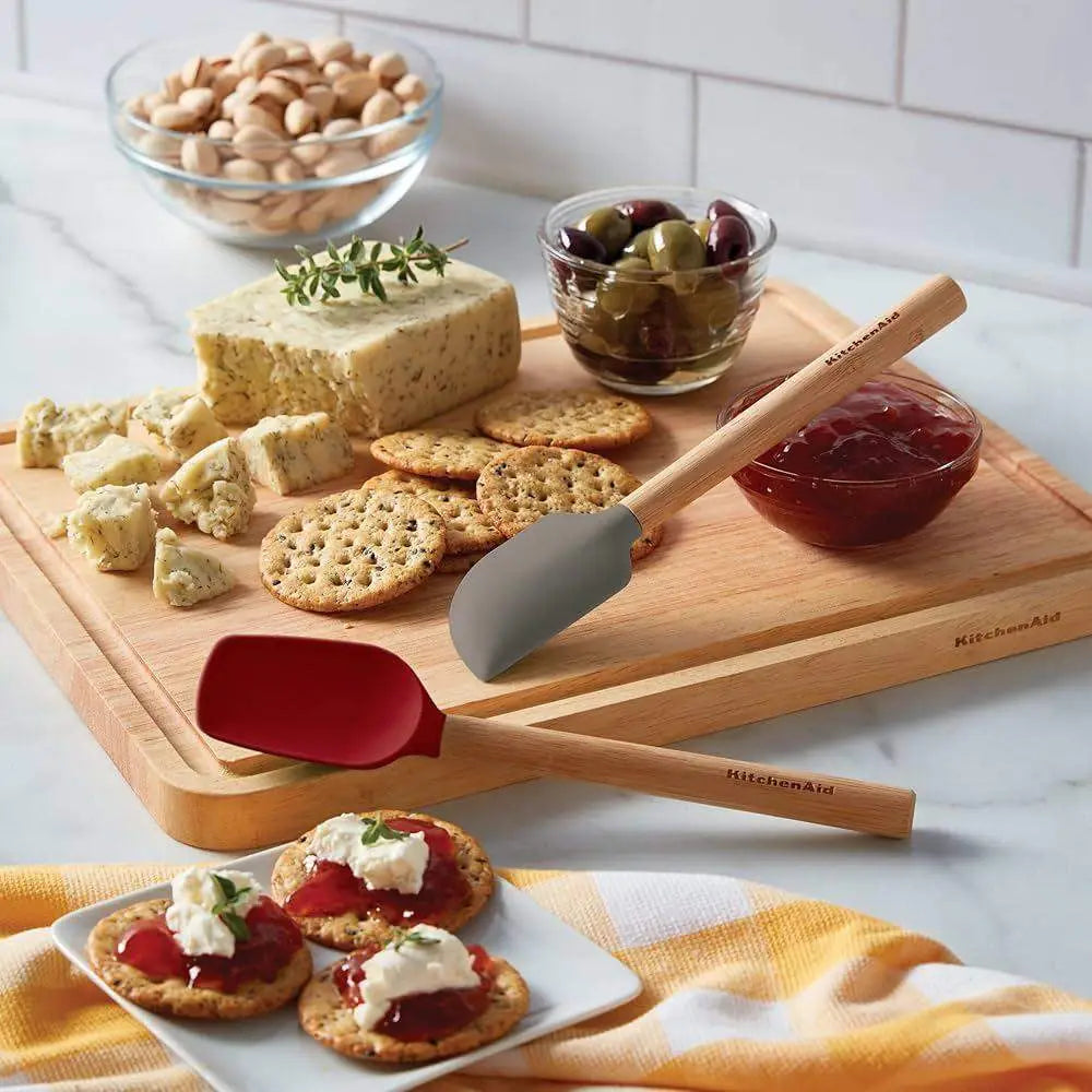 Wooden cutting board with cheese, crackers, and a bowl of olives, accompanied by KitchenAid utensils.