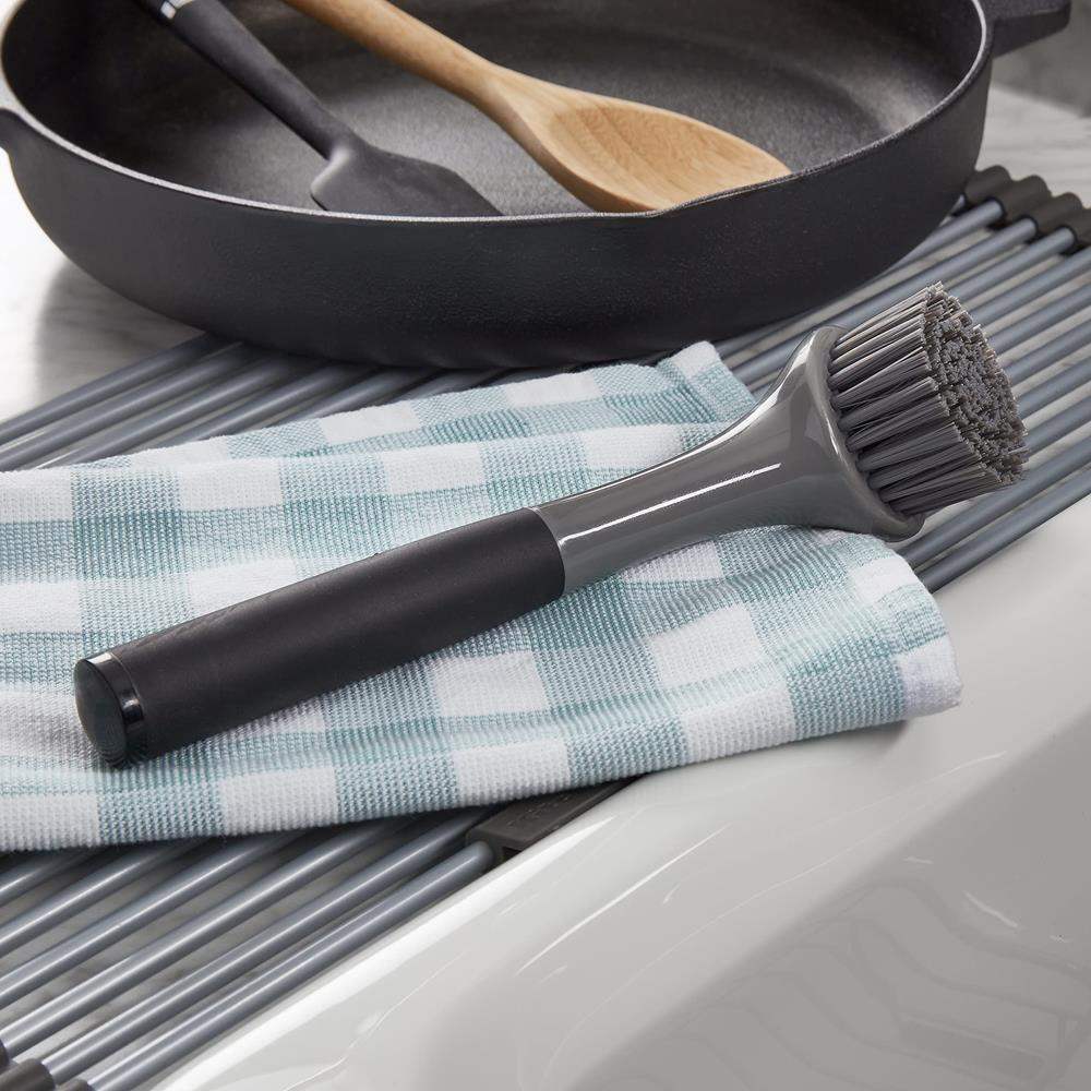 Kitchen cleaning brush with black handle and gray bristles on a checkered towel next to a frying pan.