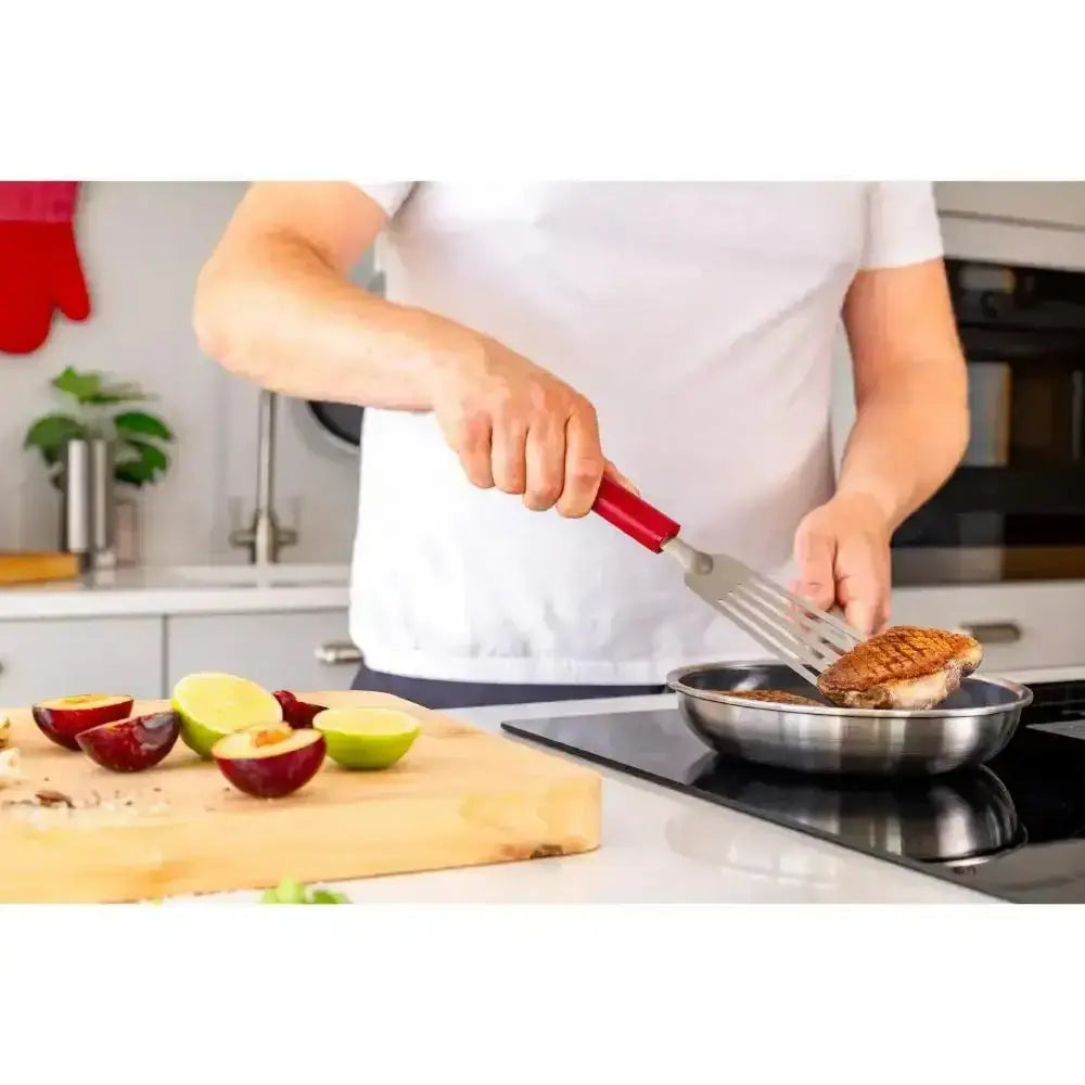 Person cooking a steak on a stove with a spatula, surrounded by fruits on a cutting board.