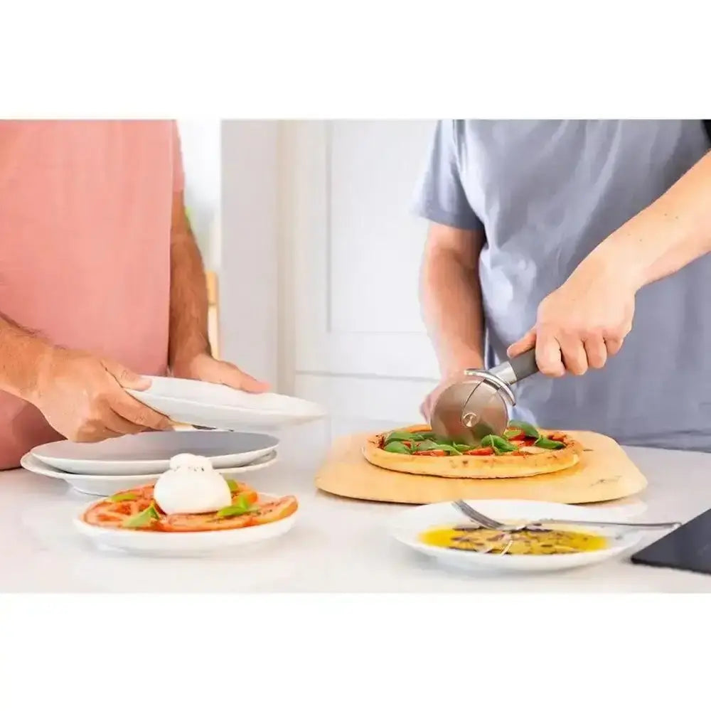 Person cutting a pizza with a pizza cutter on a table with plates and utensils.