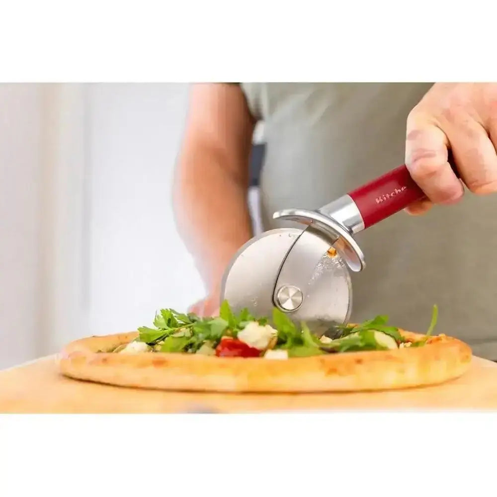 Person using a red pizza cutter on a pizza with various toppings on a wooden board.