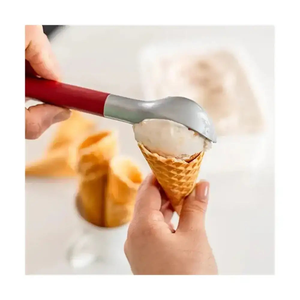 Hand holding a waffle cone with ice cream being scooped into it using a red-handled ice cream scoop.