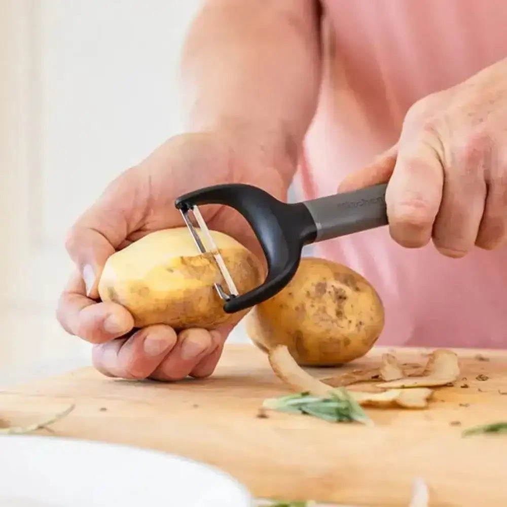Person peeling a potato with a black and grey peeler on a wooden cutting board.