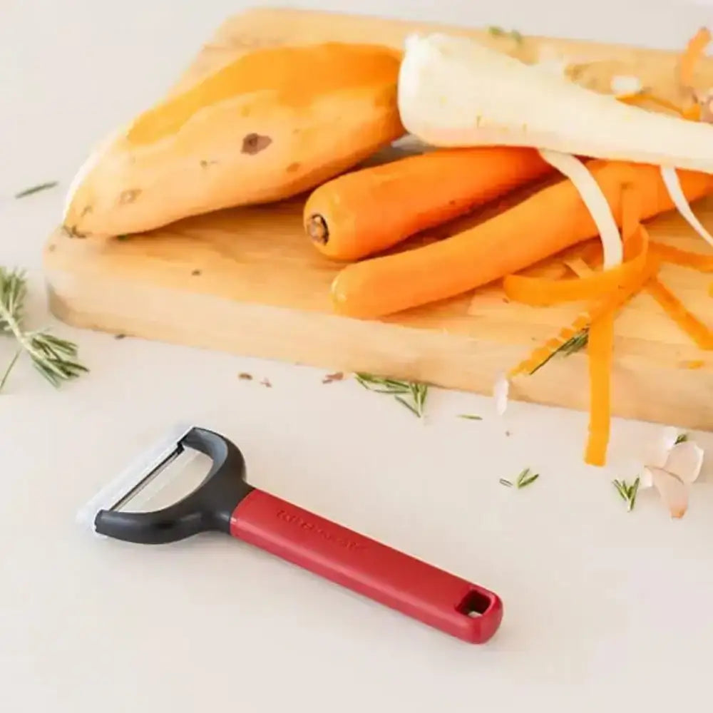 Red vegetable peeler on a white surface with carrots and sweet potatoes on a wooden cutting board.