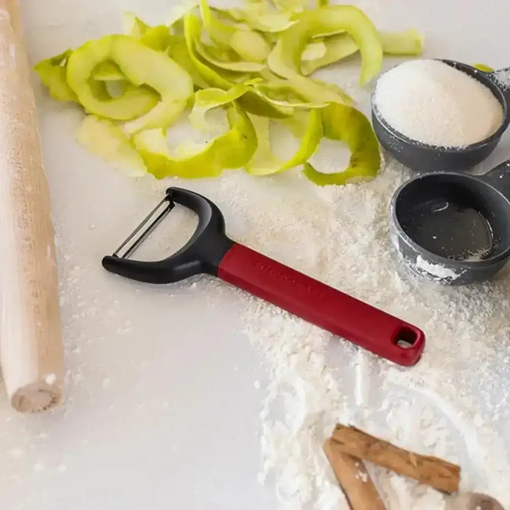 Red and black vegetable peeler on a white surface with sliced green apples, a rolling pin, and small bowls.