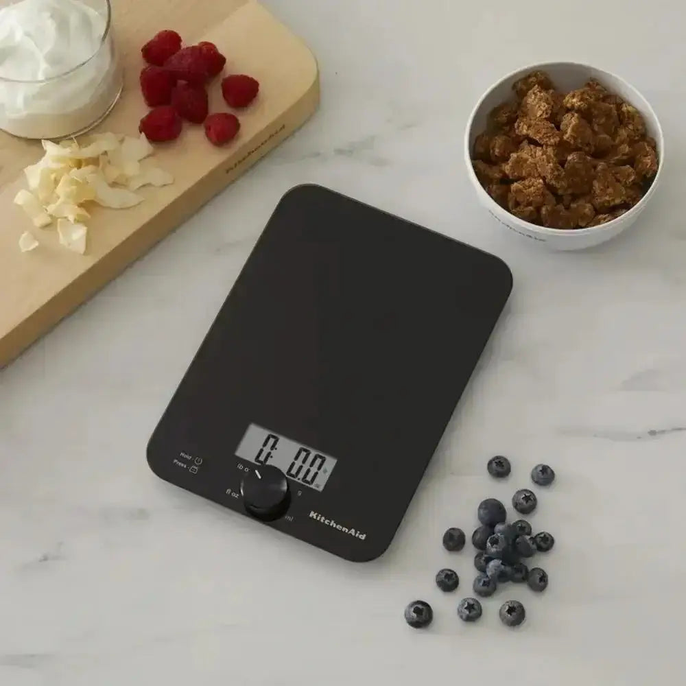 Black digital kitchen scale on a marble surface with fruits and a bowl of brown sugar.