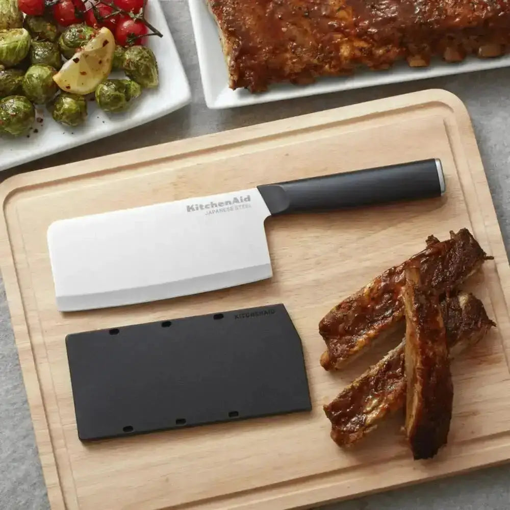 KitchenAid cleaver on a wooden cutting board with cooked ribs and vegetables in the background.