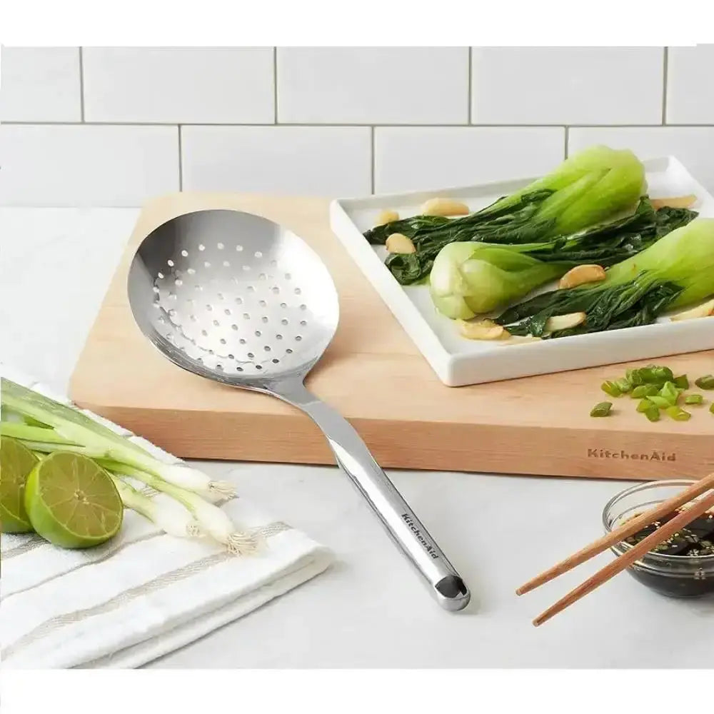 Stainless steel slotted spoon on a cutting board with vegetables and kitchen items.