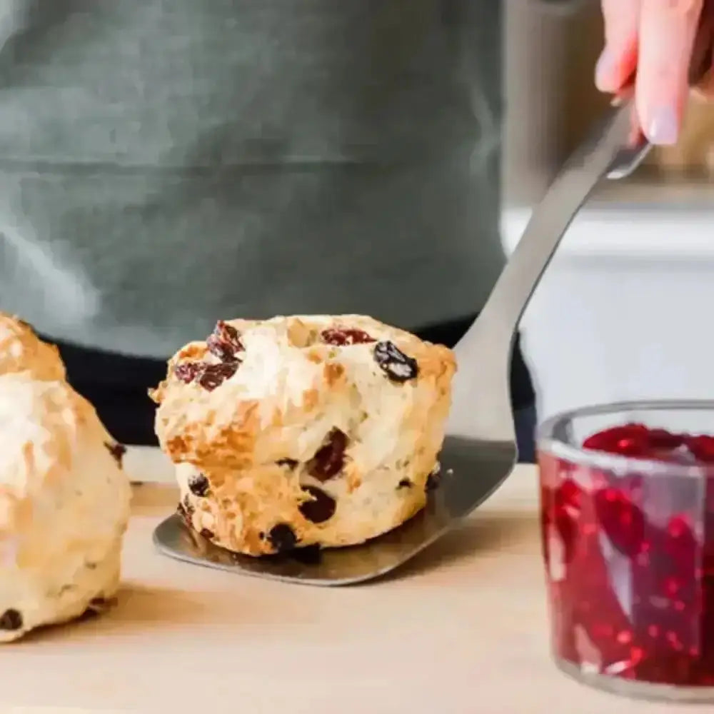 Muffin with raisins being topped with red jam on a wooden surface.