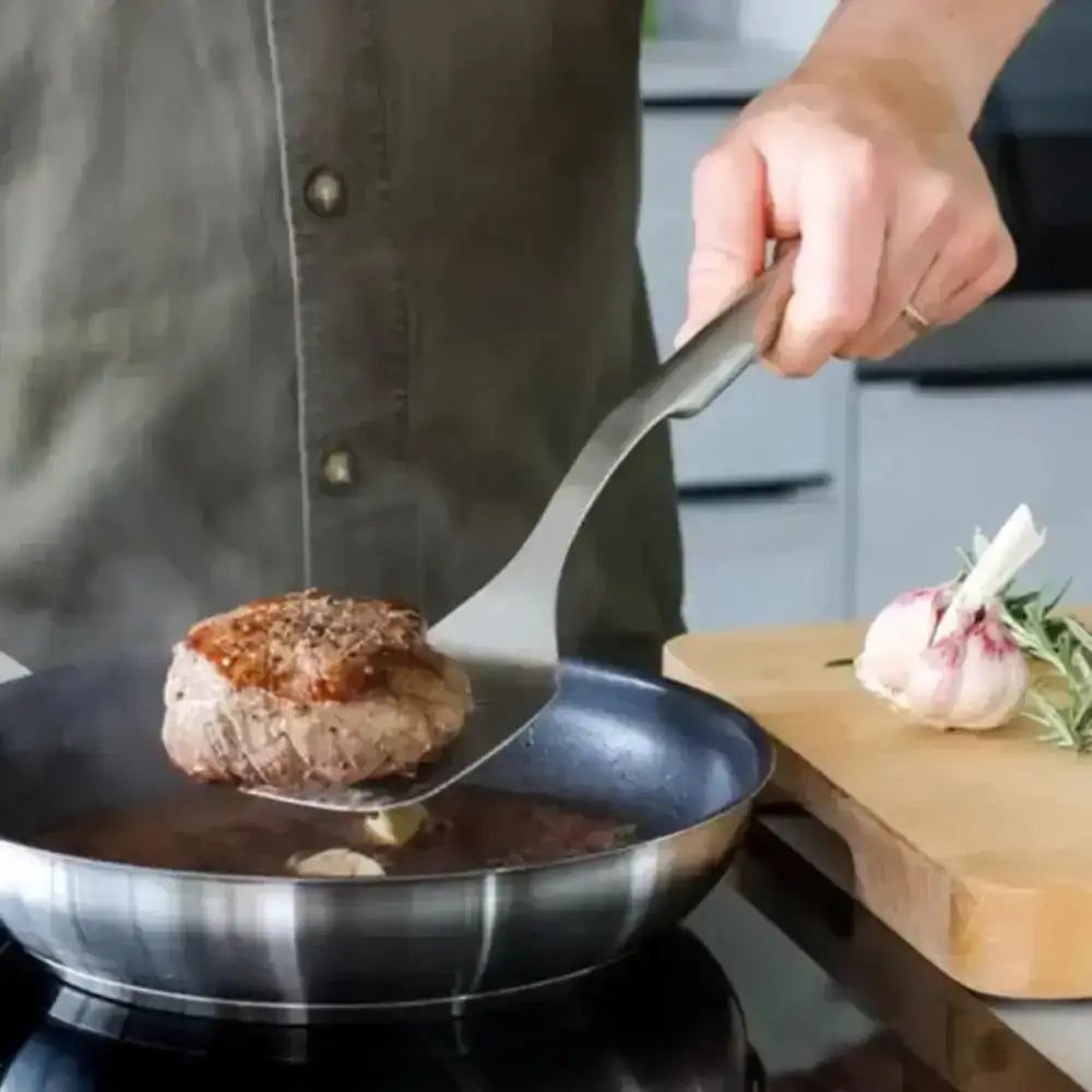 Person cooking a meat patty in a pan with a wooden cutting board in the background