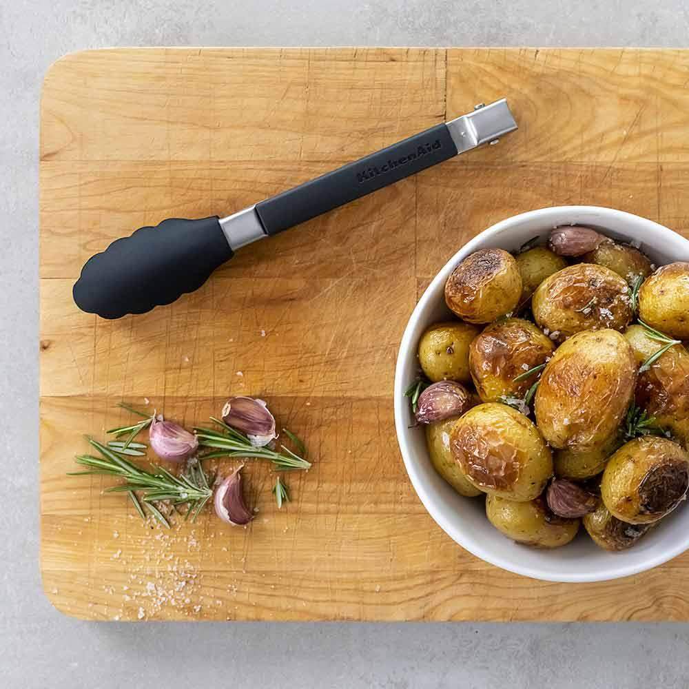 Wooden cutting board with roasted potatoes, rosemary, garlic, and tongs on a gray surface.